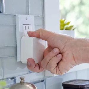 Hand plugging a white smart home device into a wall outlet in a kitchen.