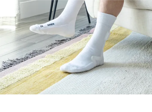 Person sitting on a couch wearing white socks with the word 'Feetwings' and an 'R' on the right sock, resting feet on a multi-colored rug.