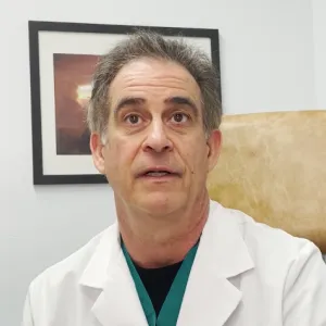 Middle-aged male doctor in a white coat sitting in an office with framed artwork on the wall behind him.