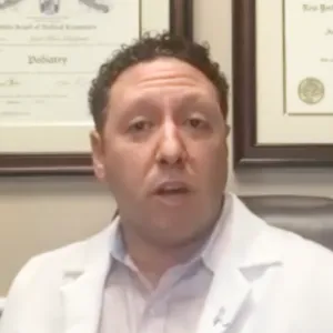 Man with curly hair wearing a white medical coat sitting in front of framed certificates on a wall.