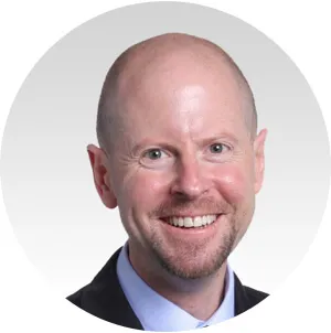 Smiling bald man with light complexion wearing a dark suit jacket and light blue shirt against a white background.