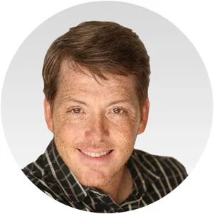 Smiling man with short brown hair and freckles wearing a dark plaid shirt against a light gray background.