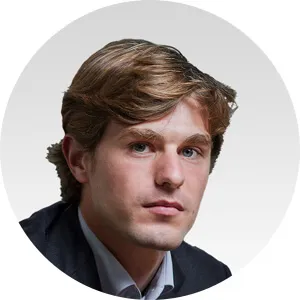 Young man with light brown hair wearing a dark blazer and light shirt against a light gray circular background.