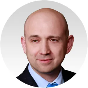 Portrait of a bald man wearing a dark suit jacket, light blue shirt, and blue tie against a light gray background.