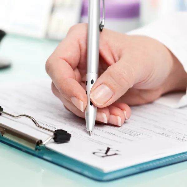 Hand holding a silver pen poised to sign a prescription form on a blue clipboard.