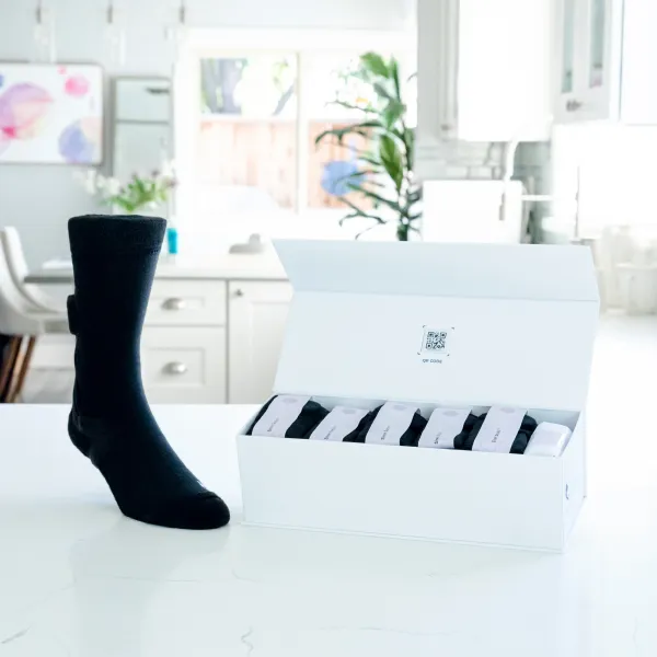 Black sock standing upright next to an open white box containing six pairs of black socks on a white kitchen counter.