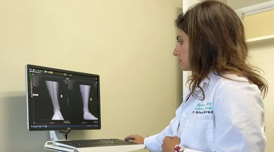 Medical professional in a white lab coat examining X-ray images of a lower leg displayed on a computer monitor.