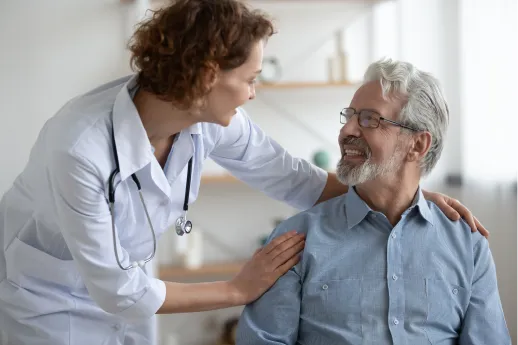 Doctor in white coat with stethoscope smiling and talking to an older male patient who is sitting and smiling back.